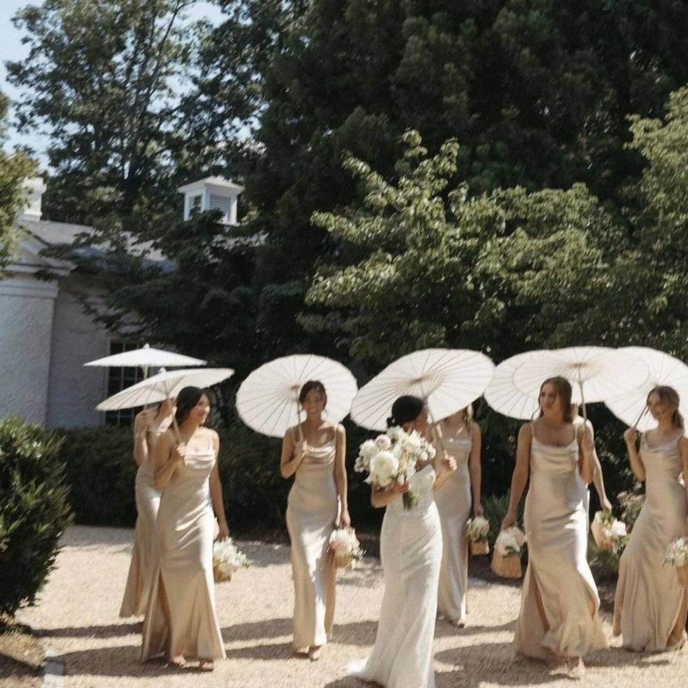 The Full Party package: bride and bridesmaids in champagne silk dresses walking a sunlit garden path while holding white paper parasols and ivory bouquets
